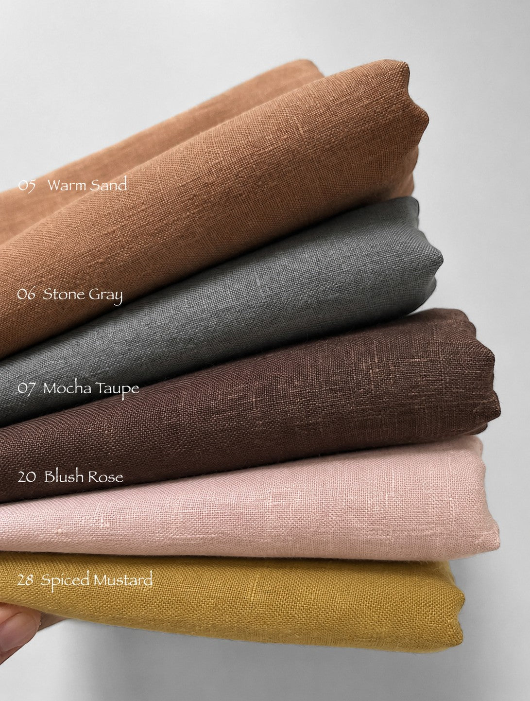 Six folded fabric swatches stacked diagonally on a light gray background, with text labels identifying the colors: Warm Sand, Stone Gray, Mocha Taupe, Blush Rose, and Spiced Mustard. A hand is visible at the bottom left holding the stack.